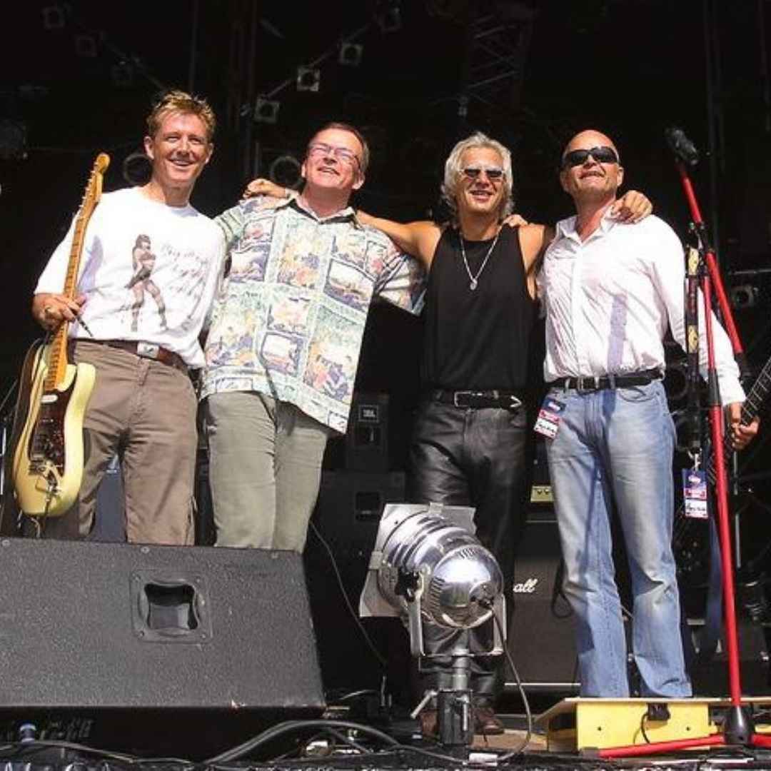 Flangia Kaiphos stehen zusammen auf der Bühne, jeder trägt unterschiedliche Kleidung und hält Instrumente.